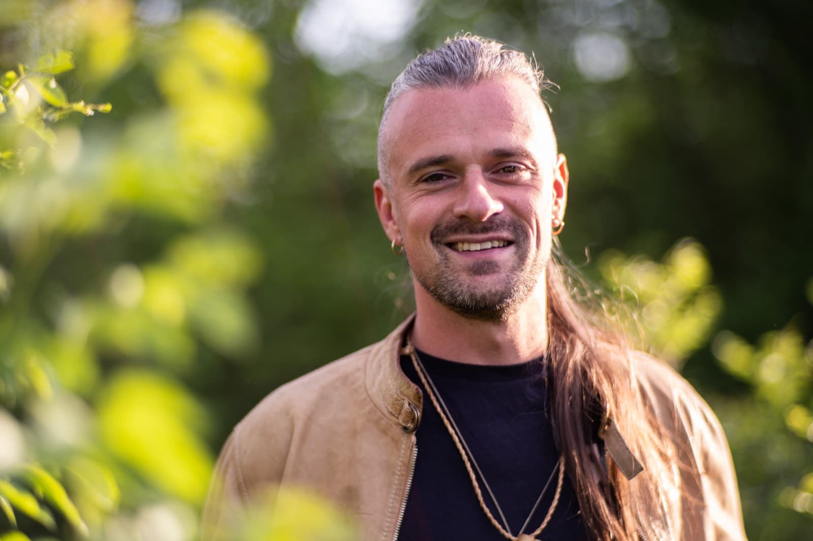 Portrait of Axel Drioli smiling, in a tan jacket against green woodland